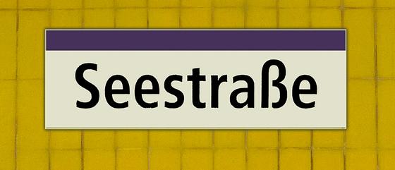 U-Bahnhof Seestraße Kaffeetasse - schwarze Schrift auf weißem Schild mit lila Streifen, vor gelber Mosaikwand