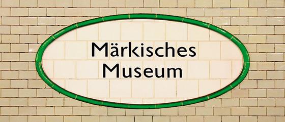 U-Bahnhof Märkisches Museum Kaffeetasse - schwarze Schrift auf ovalem, weißem Schild aus Fliesen, mit grüner Umrandung, vor hellbeigen Kacheln