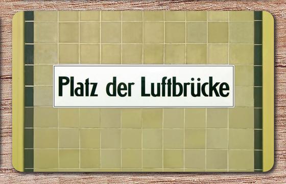 U-Bahnhof Platz der Luftbrücke Frühstücksbrettchen - schwarze Schrift auf weißem Schild, vor beigen Mosaikfliesen mit schwarzem Streifen rechts und links