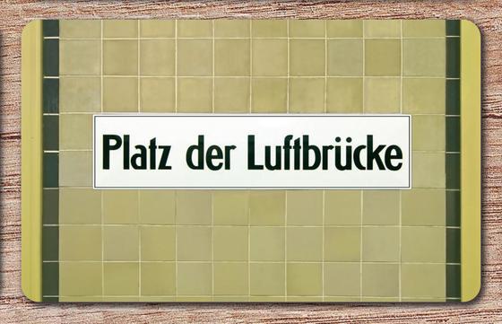 U-Bahnhof Platz der Luftbrücke Frühstücksbrettchen - schwarze Schrift auf weißem Schild, vor beigen Mosaikfliesen mit schwarzem Streifen rechts und links