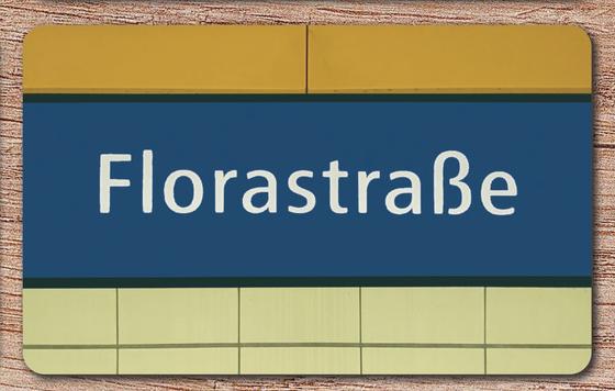 U-Bahnhof Florastraße Früstücksbrettchen - weiße Schrift auf blauem Schild, zwischen gelbem und beigen Platten