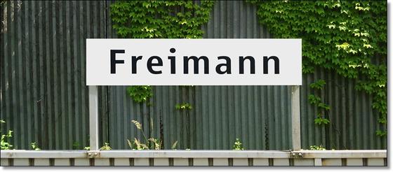 U-Bahnhof Freimann Kaffeetasse - schwarze Schrift auf weißem Schild vor grauem Wellblech mit Rankpflanze