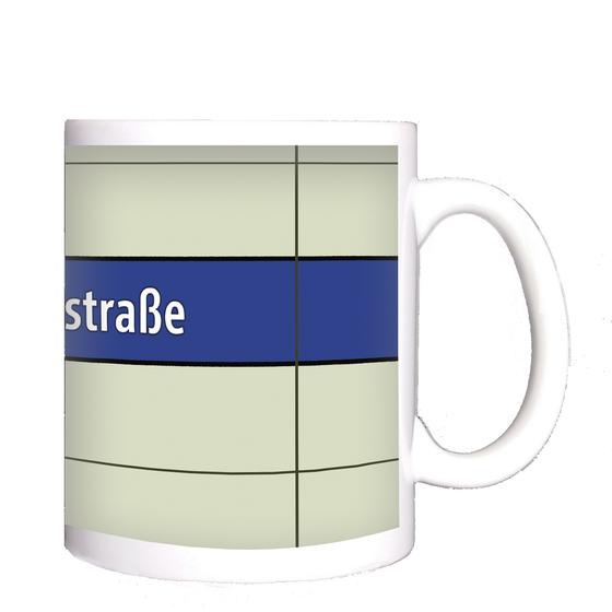 U-Bahnhof Giselastraße Kaffeetasse - weiße Schrift auf blauem Streifen, vor weißem Plattenhintergrund