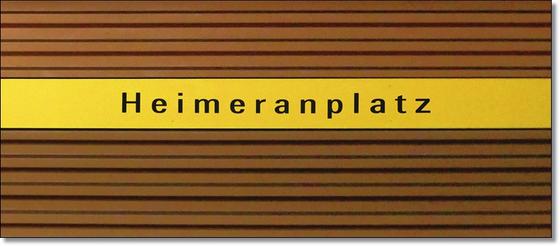 U-Bahnhof Heimeranplatz Kaffeetasse - schwarze Schrift auf gelbem Streifen, vor braunem Riffelhintergrund