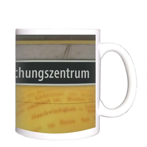 U-Bahnhof Garching-Forschungszentrum Kaffeetasse - weiße Schrift auf schwarzem Schild, zwischen gelblicher Plattenwand mit Buchseiten-Print und grauen Platten