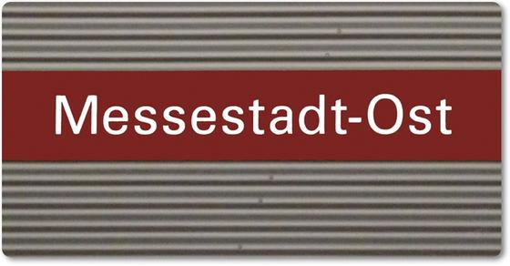 U-Bahnhof Messestadt-Ost Kühlschrankmagnet - weiße Schrift auf dunkelrotem Streifen, vor grauer Wellblechwand