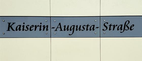 U-Bahnhof Kaiserin-Augusta-Straße Kaffeetasse - Schwarze, geschwungene Schrift auf grau-blauem Schild vor weißem Fliesenhintergrund