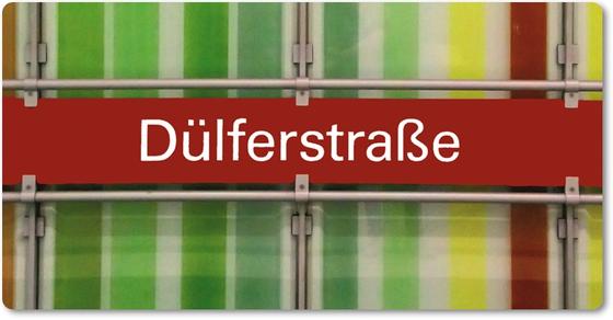 U-Bahnhof Dülferstraße Kühlschrankmagnet- weiße Schrift auf rotem Schild auf Metallgestell, vor Streifenhintergrund in diversen Grüntönen