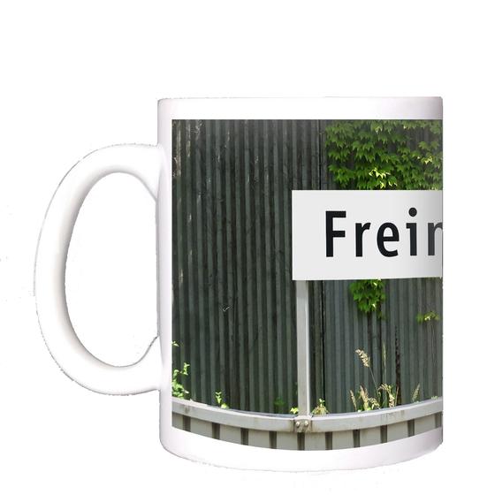 U-Bahnhof Freimann Kaffeetasse - schwarze Schrift auf weißem Schild vor grauem Wellblech mit Rankpflanze