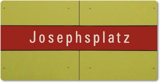 U-Bahnhof Josephsplatz - weiße Schrift auf dunkelrotem Streifen vor grünlich gelber Wand