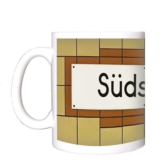 U-Bahnhof Südstern Kaffeetasse - weißes Schild mit schwarzer, schlichter Schrift auf weißem Schild mit rötlich-brauner Umrandung auf orange-gelben Fliesen
