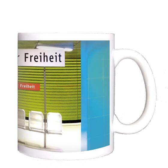 U-Bahnhof Münchner-Freiheit Kaffeetasse - weißes Schild mit schwarzer Schrift, im Hintergrund der Bahnhof mit zwei blauen Säulen, einer grasgrünen Wand in Jalousieoptik und fünf silbernen Bahnhofssitzen