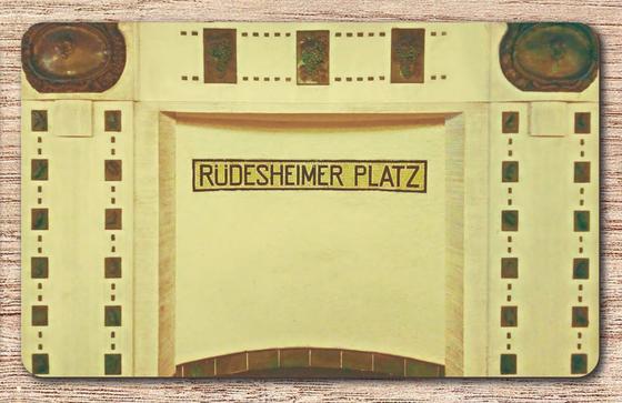 U-Bahnhof Ruedesheimer Platz Frühstücksbrettchen - schwarzer Schrift auf gelbem Mosaikstreifen, auf beiger Wand mit brauner, detaillierter Verzierung