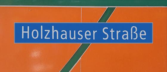 U-Bahnhof Holzhauser Straße Kaffeetasse - weiße Schrift auf blauem Schild vor orangem Hintergrund mit dunkelgrünem, diagonalem Streifen