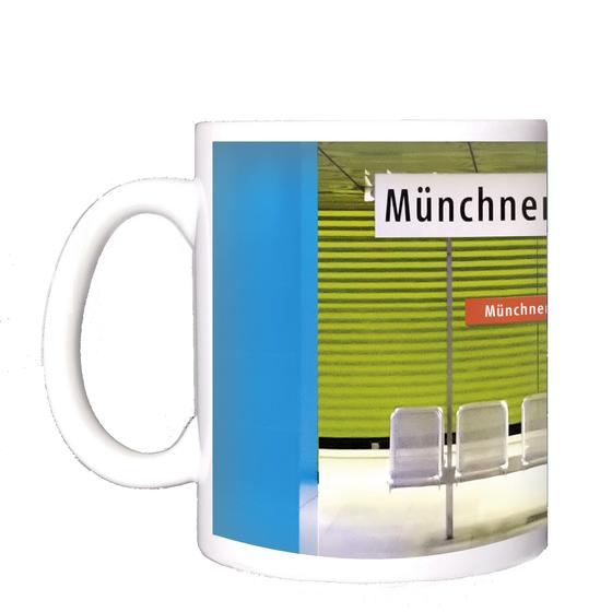 U-Bahnhof Münchner-Freiheit Kaffeetasse - weißes Schild mit schwarzer Schrift, im Hintergrund der Bahnhof mit zwei blauen Säulen, einer grasgrünen Wand in Jalousieoptik und fünf silbernen Bahnhofssitzen