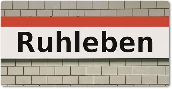 U-Bahnhof Ruhleben- schwarze Schrift auf weißem Schild mit orangem Streifen auf weißer Fliesenwand