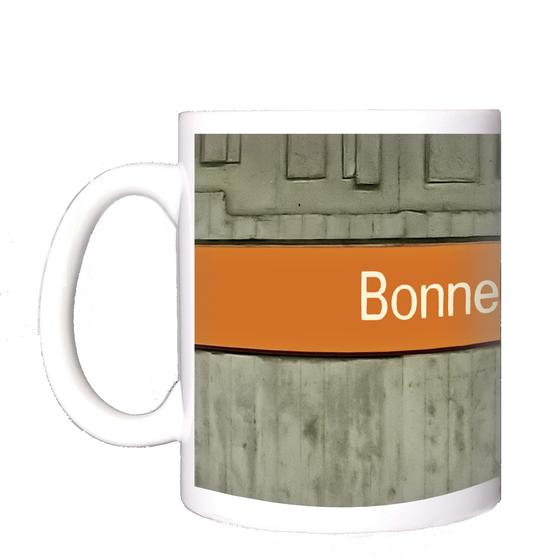 U-Bahnhof Bonner Platz Kaffeetasse - weiße Schrift auf orangem Streifen, vor grauer Betonwand