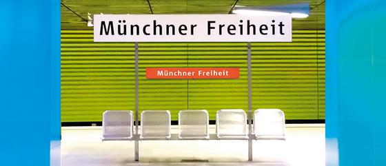 U-Bahnhof Münchner-Freiheit Kaffeetasse - weißes Schild mit schwarzer Schrift, im Hintergrund der Bahnhof mit zwei blauen Säulen, einer grasgrünen Wand in Jalousieoptik und fünf silbernen Bahnhofssitzen