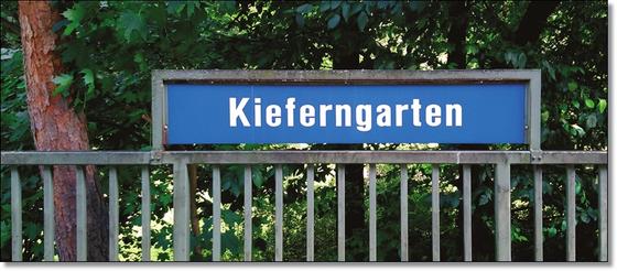 U-Bahnhof Kieferngarten Kaffeetasse - weiße Schrift auf blauem Schild, über Metallzaun, vor Bäumen