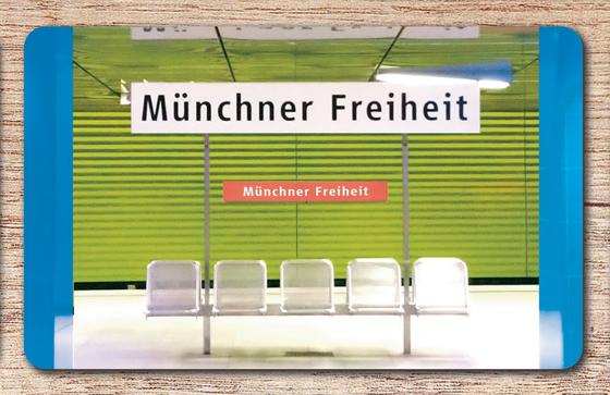 U-Bahnhof Münchner-Freiheit Frühstücksbrettchen - weißes Schild mit schwarzer Schrift, im Hintergrund der Bahnhof mit zwei blauen Säulen, einer grasgrünen Wand in jalousieoptik und fünf silbernen Bahnhofssitzen