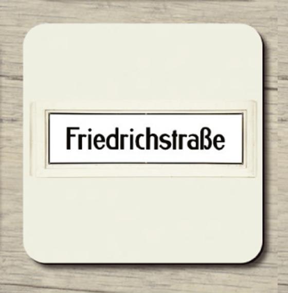 U-Bahnhof Friedrichstraße - schwarze Schrift auf weißem Schild mit schwarzer Umrandung und beige Wand im Hintergrund 