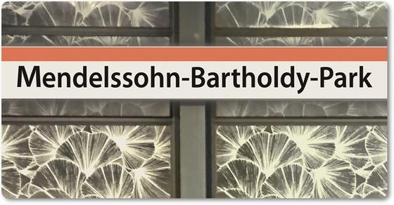 U-Bahnhof Mendelssohn-Bartholdy-Park- schwarze Schrift auf weißem Schild mit orangem Streifen vor weißem Kratzmuster in Blumenoptik