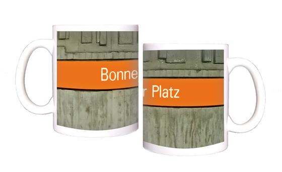 U-Bahnhof Bonner Platz Kaffeetasse - weiße Schrift auf orangem Streifen, vor grauer Betonwand