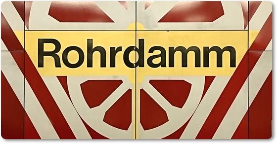U-Bahn-Magnet Berlin Rohrdamm - schwarze Schrift auf gelbem Schild, umrandet von weißem, radähnlichem Muster auf rot-braunem Untergrund