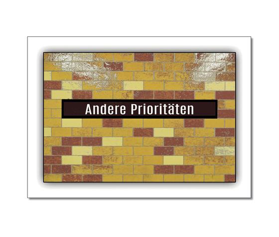 Postkarte in Ubahnstationsschild-Optik - laengliche klinkerartige Fliesen in horizontal versetzter Anordnung, überwiegend Orange, partziell blass gelb und dunkles Rot, darauf zentral auf schwarzem Schild in weisser Schrift "Andere Prioritaeten"