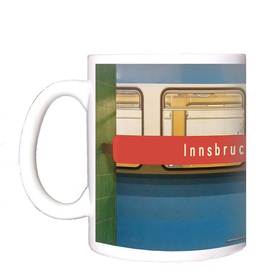 U-Bahnhof Innsbrucker Ring Kaffeetasse - weiße Schrift auf rotem Streifen, vor blauem U-Bahnwagon, zwischen grünen Säulen
