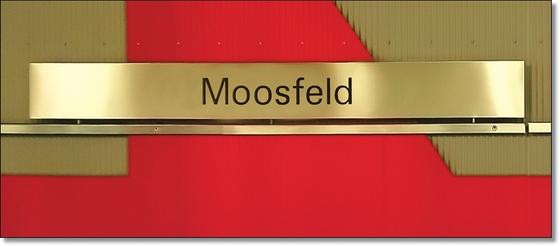 U-Bahnhof Moosfeld Kaffeetasse - schwarze Schrift auf goldenem Schild, vor rotem Untergrund mit grünlichen Elementen
