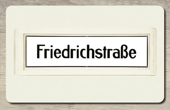 U-Bahnhof Friedrichstraße - schwarze Schrift auf weißem Schild mit schwarzer Umrandung und beige Wand im Hintergrund 