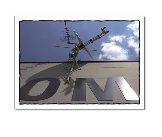 Klassische Postkarte - Foto einer Anbindung an einer Hauswand. Auf der Hauswand stehen die Buchstaben O und M unter einer Satellitenschuessel. 