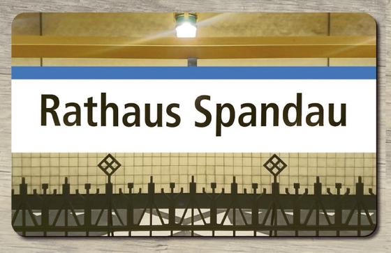Endstation U-Bahnhof Rathaus Spandau - gusseiserne verschnoerkelte Zaunelemente, im Hintergrund kleine helle Fliesen, auf dem Zaun quer übers Bild der Schriftzug in schwarz auf weissem Untergrund mit dem blauen Farbbalken der Linie U7 oberhalb der Schrift