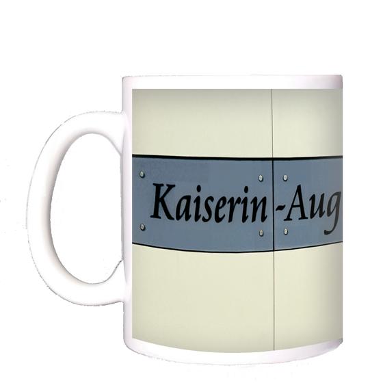 U-Bahnhof Kaiserin-Augusta-Straße Kaffeetasse - Schwarze, geschwungene Schrift auf grau-blauem Schild vor weißem Fliesenhintergrund