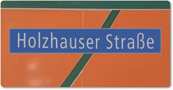 U-Bahnhof Holzhauser Straße Magnet - weiße Schrift auf blauem Schild vor orangem Hintergrund mit dunkelgrünem, diagonalem Streifen