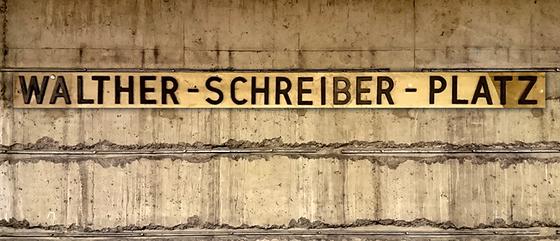 U-Bahnhof Walther-Schreiber-Platz Kaffeetasse - ehemalige Stationsgestaltung, schwarze Schrift auf Messingschild vor grau-brauner Betonwand