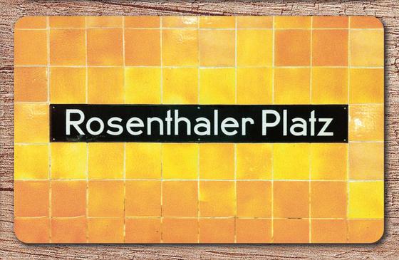U-Bahnhof Rosenthaler Platz Frühstücksbrettchen - schwarzes Schild mit weißer Schrift auf orange-gelbem Fliesenhintergrund
