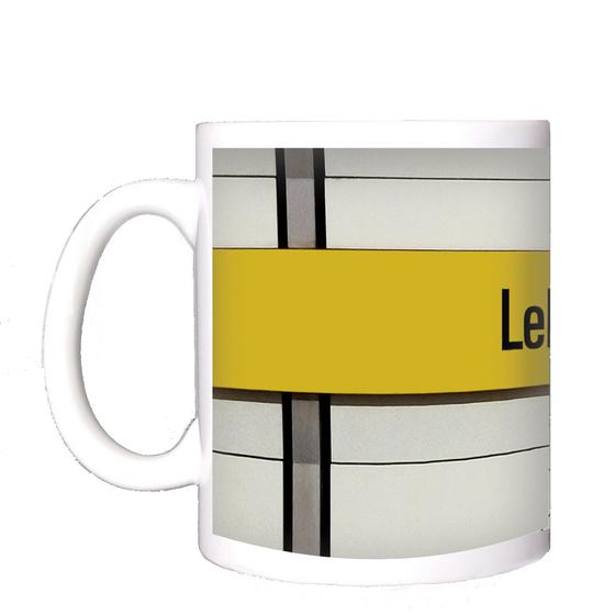 U-Bahnhof Lehel Kaffeetasse - schwarze Schrift auf gelbem Streifen, vor weißer Plattenwand