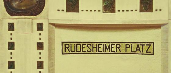 U-Bahnhof - Rüdesheimer Platz - schwarze Schrift und ein helles Schild mit schwarzem Rand (aus Mosaik) in einer Wandnische weitere geografische Verzierungen - Ansicht links und rechts