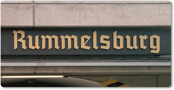 S-Bahnhof Rummelsburg - Frakturschrift in beige auf einem dunklen Schild weitere helle Streifen