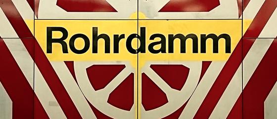 U-Bahn-Kaffeetasse Berlin Rohrdamm - schwarze Schrift auf gelbem Schild, umrandet von weißem, radähnlichem Muster auf rot-braunem Untergrund