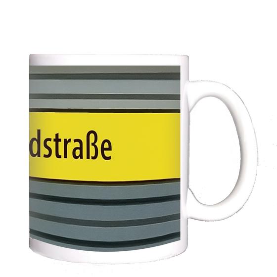 U-Bahnhof Westendstraße Kaffeetasse - schwarzer Schrift auf gelbem Schild, vor grau-blauer Wand mit schwarzen Streifen