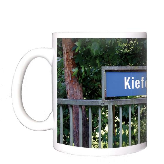 U-Bahnhof Kieferngarten Kaffeetasse - weiße Schrift auf blauem Schild, über Metallzaun, vor Bäumen