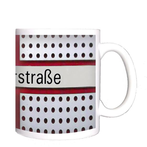 U-Bahnhof Kreillerstraße Kaffeetasse - schwarze Schrift auf weißem Schild, welches Teil eines roten Kreuzes ist, vor zweigeteiltem Hintergrund mit schwarz-weißen Punkten und rot-braunem Backsteinmuster