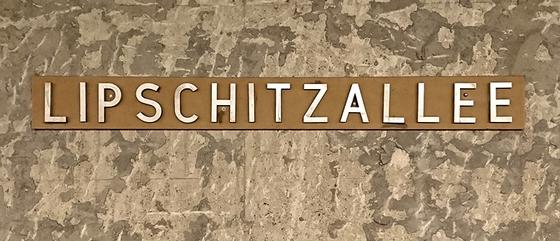 U-Bahnhof Lipschitzallee Kaffeetasse - weiße Schrift auf bronzefarbenem Schild, vor beige-braun geflecktem Hintergrund