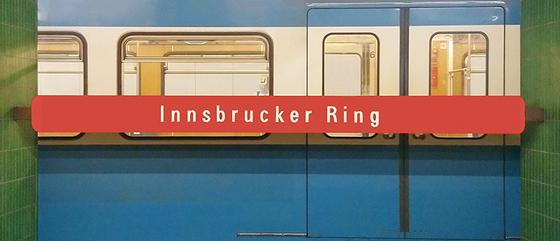 U-Bahnhof Innsbrucker Ring Kaffeetasse - weiße Schrift auf rotem Streifen, vor blauem U-Bahnwagon, zwischen grünen Säulen