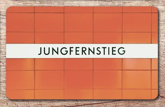 U-Bahnhof Jungfernstieg Frühstücksbrettchen - schwarze Schrift auf weißem Streifen vor oranger Fliesenwand