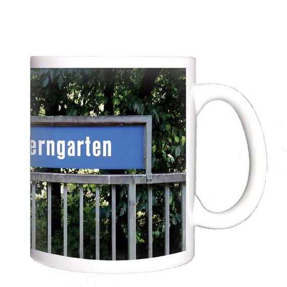 U-Bahnhof Kieferngarten Kaffeetasse - weiße Schrift auf blauem Schild, über Metallzaun, vor Bäumen