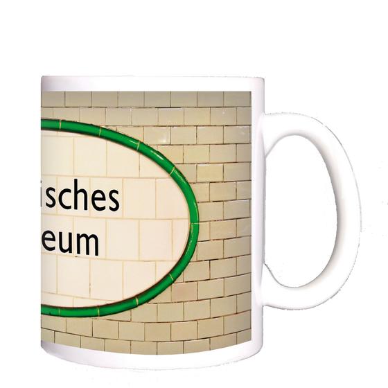 U-Bahnhof Märkisches Museum Kaffeetasse - schwarze Schrift auf ovalem, weißem Schild aus Fliesen, mit grüner Umrandung, vor hellbeigen Kacheln